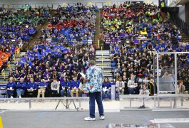 Man in blue shirt standing at podium in front of crowd of people.