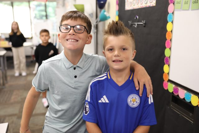 two elementary age boys standing next to each other boy in gray shirt with glasses has arm around boy with blue shirt