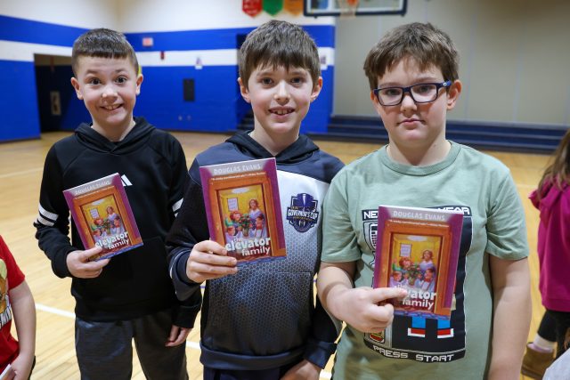 Three young boys proudly holding books in a gymnasium.