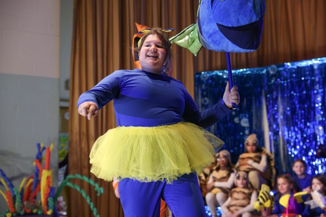 A child in a blue and yellow costume, dressed as Dory from FInding Nemo, standing on a stage with a microphone, smiling and