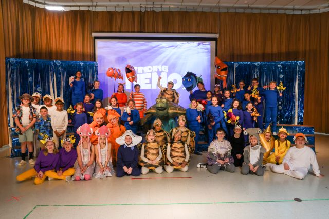 The cast of Finding Nemo in costumes, posing for a photo on a stage with a backdrop of Finding Nemo sign