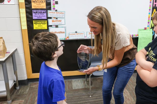 Woman in a brown shirt holding an award, standing between two children in a classroom setting.