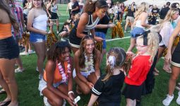 A group of cheerleaders, high school students, and younger students in a stadium setting, with some standing and others sitting on the grass talking and trading pom-poms.
