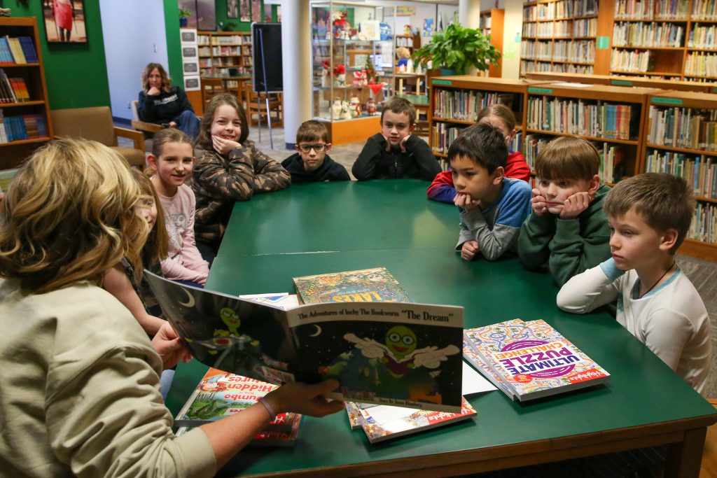 Children gathered around a table in a library setting, listening to an adult reading a book aloud.
