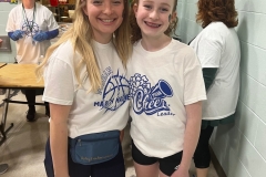 Two women posing in a school gymnasium, one wearing a white t-shirt and the other a blue headband, both smiling at the