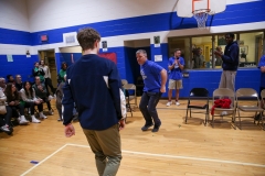 People in a gymnasium playing basketball, one person is standing on the court while others are seated around the room.