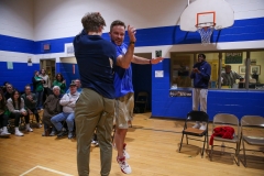 Two men playing basketball in a gymnasium, one jumping to catch the ball while the other tries to block him.