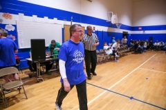 Man in blue shirt walking through gymnasium during basketball game.