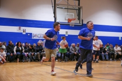 Two men in blue shirts running on a basketball court during an indoor game, one man is wearing shorts and the other is