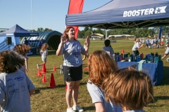 Woman in blue shirt standing at a booth with several people around her, possibly at an outdoor event like a fun run.