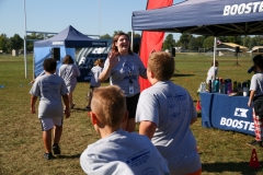 People gathered around a tent at an outdoor event, possibly a fun run, with a woman speaking to them.