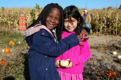 Two children posing at a pumpkin patch, one holding a pumpkin.