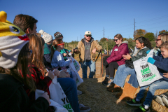 People gathered around a man in a field, some holding signs, possibly for an event or demonstration.