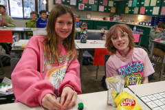 Two young girls sitting at a table in what appears to be a classroom, smiling and posing for the camera.
