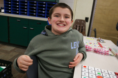 Boy in a classroom chair, smiling at camera, seated at a table with educational materials.
