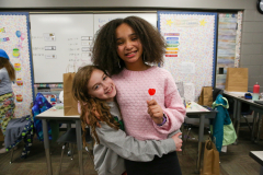 Two young girls hugging in a classroom, one holding a heart-shaped sign.