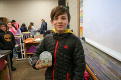 Boy holding a green frog figurine in front of a classroom setting with students in background.