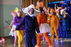 A group of people, dressed in colorful costumes, are performing in a school gymnasium.