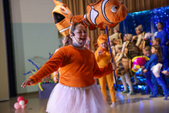 A young girl dressed as Nemo, holding a fish balloon, stands on stage at a school event.