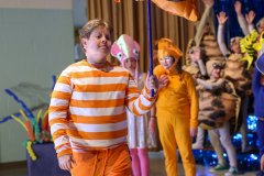 A person in an orange and white striped jumpsuit stands in front of a group of children in costumes.