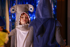 Two people in costumes, one with a shark head, the other in a hoodie, standing in front of a blue curtain.