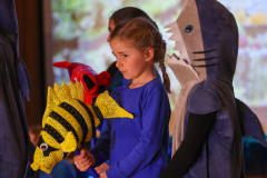 Children in costumes, one girl holding a fish-shaped toy, another child in a shark costume.