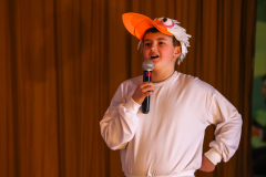 A young man in a white shirt and orange duck headband holding a microphone.