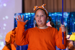 A young girl in a red shirt and orange pants is holding a blue wand and making a funny face.