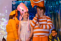 Two children dressed in costumes, one in a fish hat and the other in a clownfish outfit, standing in front of a blue curtain