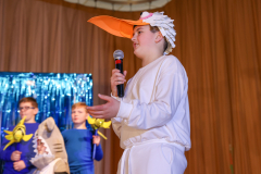 A young boy in a white robe and a large duck hat is speaking into a microphone.