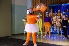 Person in a costume holding a fish, standing on a stage with a group of people dressed in costumes.