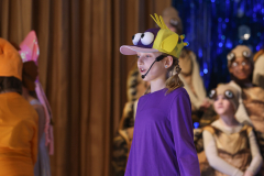 A young girl in a purple shirt and yellow hat, standing on a stage, looking to the left.