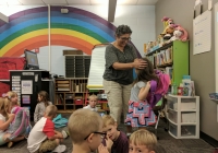 Woman standing in front of classroom desk, smiling at child with backpack.