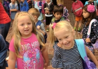 Two young girls posing in front of a brick building on their first day of kindergarten.