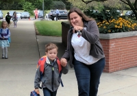 Woman and child walking together on sidewalk, both carrying backpacks, smiling at camera.