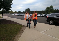 Two women walking towards camera, one holding child's hand, both wearing safety vests, in front of parked cars and parking