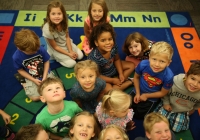 Children gathered on a colorful rug in front of an alphabet chart for their first day of kindergarten.