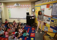 Teacher standing in front of whiteboard pointing to it, children seated on floor attentively, classroom setting, first day