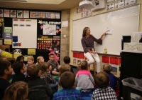 Teacher standing in front of whiteboard, writing on it, while students sit attentively at desks in classroom.