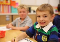 Two young boys sitting at a table in a classroom, one boy is smiling while writing on paper, the other boy is looking at the