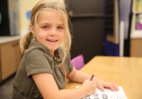 Little girl sitting at desk, writing on paper, smiling at camera.