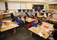 Children seated at desks in a classroom setting, engaged in an activity, possibly writing or drawing, on what appears to be