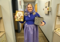 Woman in a blue dress holding a book, standing in an office setting.