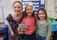 Three young girls posing in front of a classroom bulletin board with a book cover featuring cartoon characters.