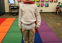 Young boy dressed as a superhero standing in front of a classroom rug, posing for a picture.