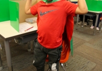 Boy dressed in a red shirt and black mask standing at a desk in a classroom.