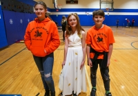 Three children posing in a gymnasium, two wearing orange shirts with white lettering and one in a white dress.