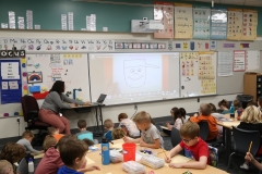Teacher presenting in front of students watching a video on a whiteboard.