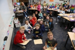 Children sitting at desks in a classroom, posing for a group photo during a pen pal meet up day.
