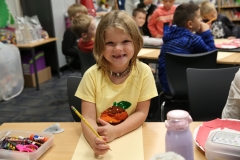 Girl in yellow shirt sitting at desk with pencil, smiling at camera.